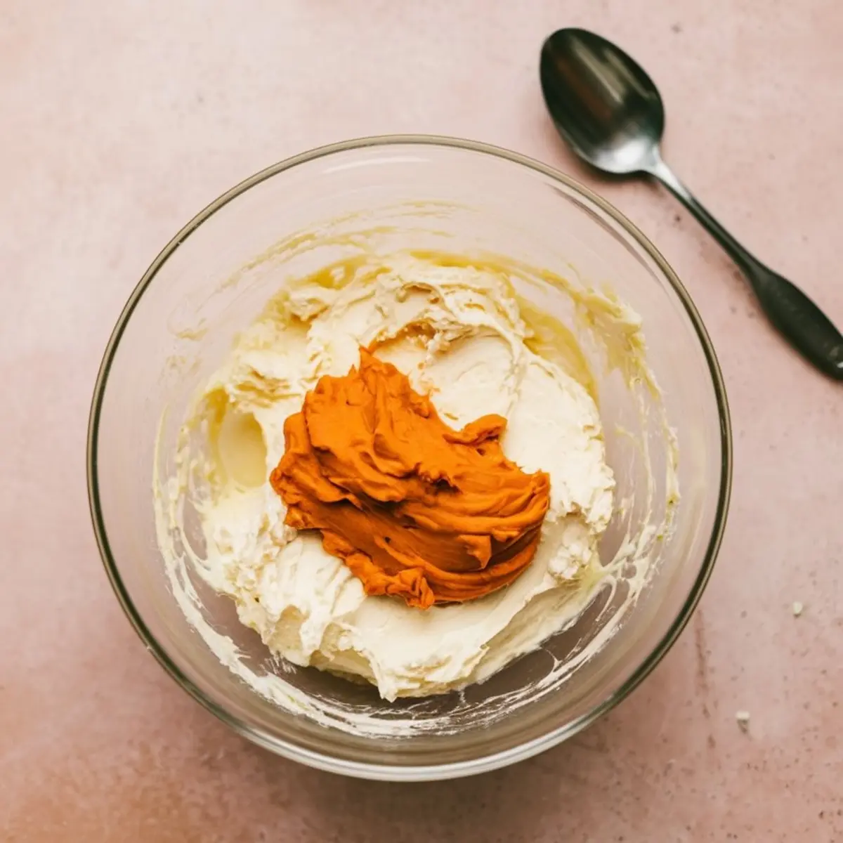 Glass bowl containing whipped cream cheese with a dollop of pumpkin puree on top, set on a pink surface with a metal spoon nearby.
