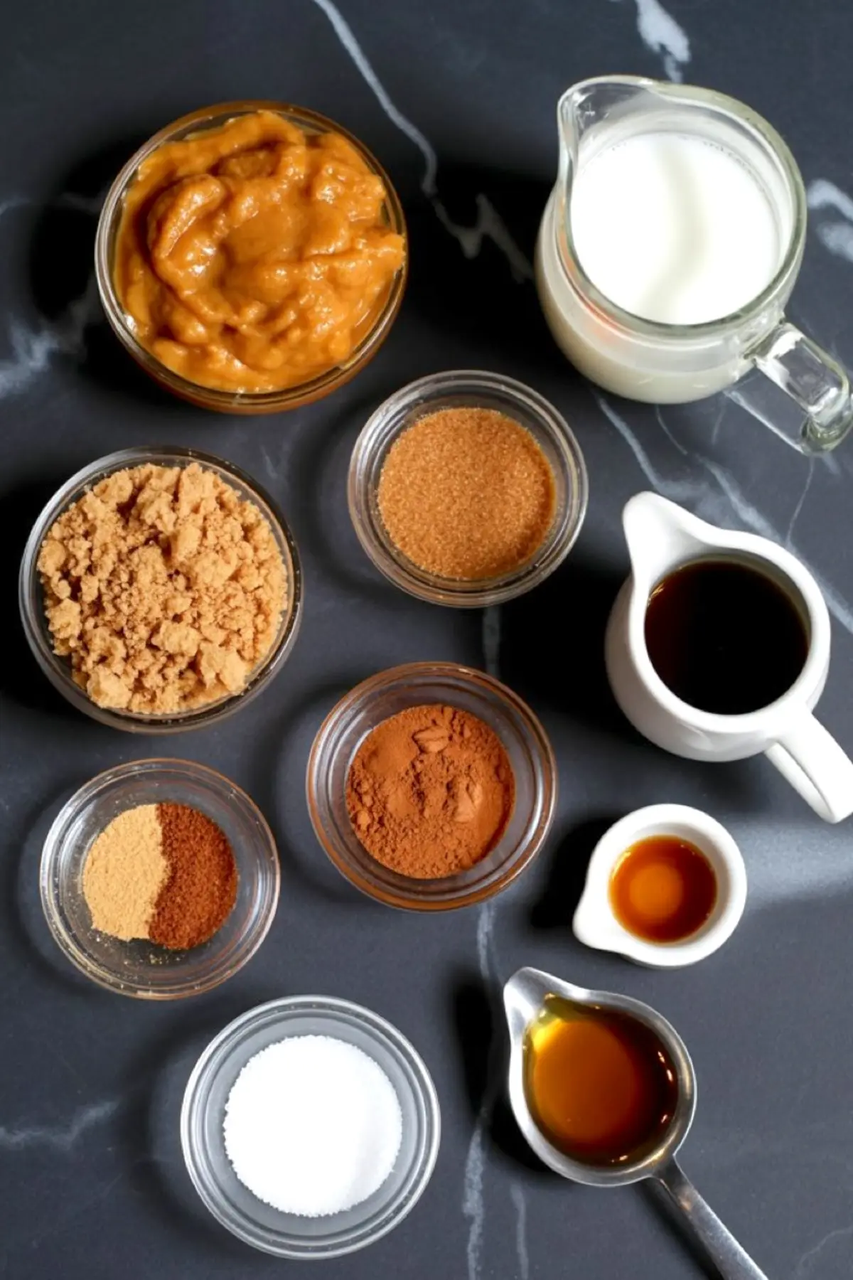 Flat lay of pumpkin spice latte ingredients including pumpkin purée, brown sugar, cinnamon, nutmeg, ginger, vanilla extract, maple syrup, milk, and brewed espresso on a dark marble background.