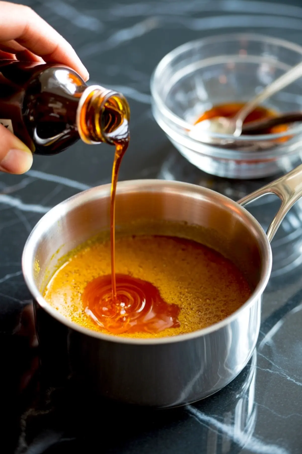 Dark vanilla syrup being poured into a saucepan filled with hot pumpkin spice latte base, with a glass bowl and spoon in the background.