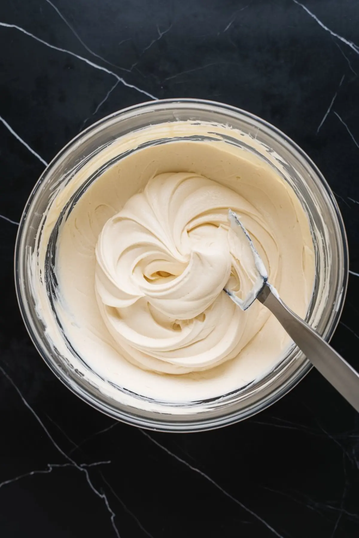Glass bowl filled with smooth cream cheese frosting for red velvet cupcakes. The frosting is swirled into soft peaks with a spatula resting on the edge of the bowl.