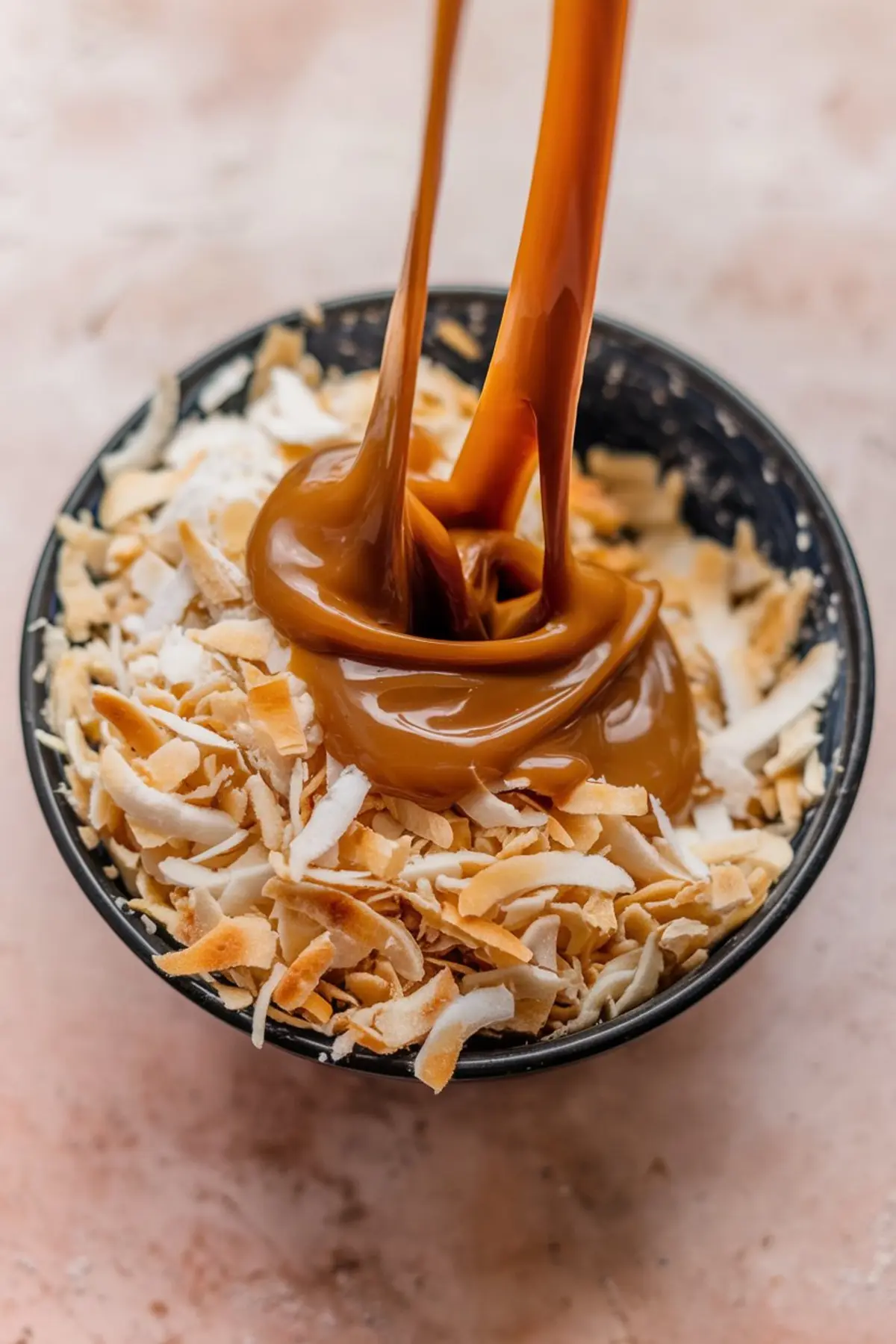 Close-up of thick caramel sauce being poured over toasted shredded coconut in a small black bowl, placed on a light pink surface.