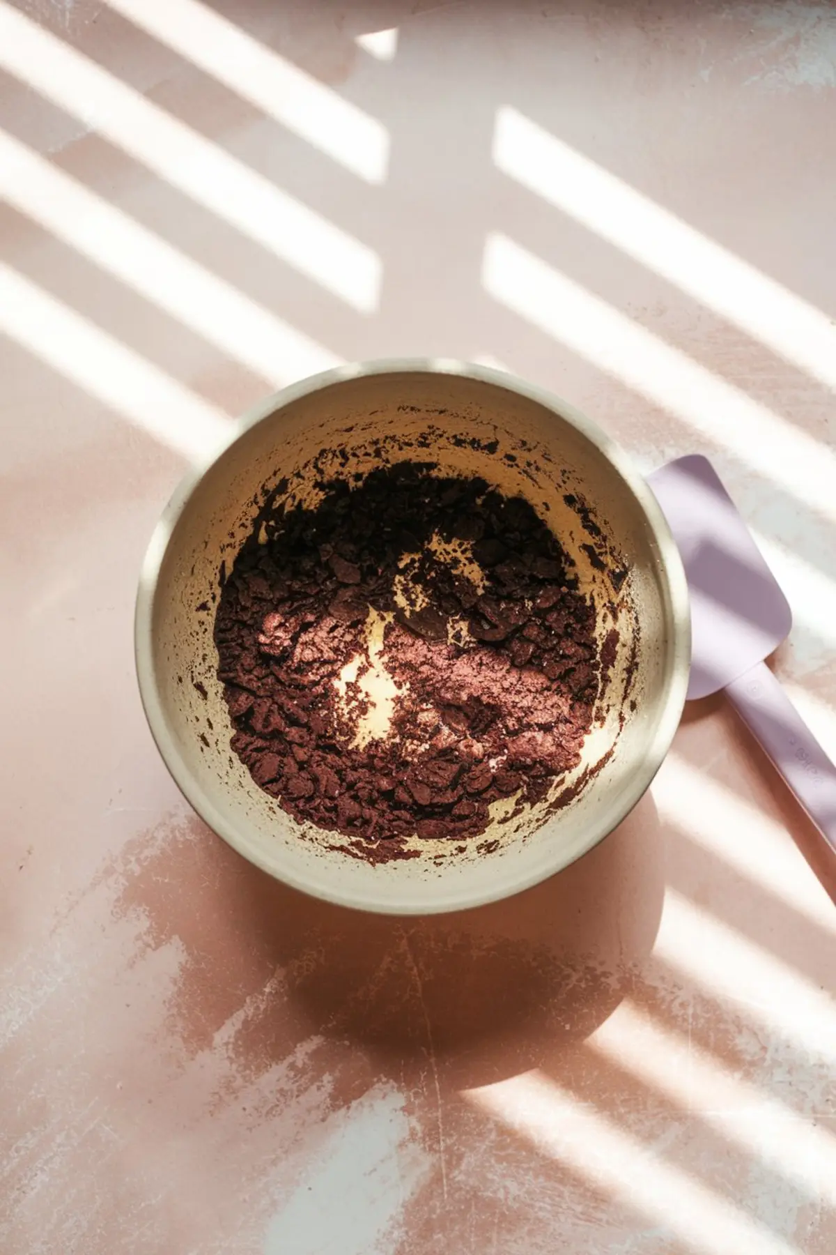 Close-up overhead shot of crushed chocolate cookies in a mixing bowl on a pink surface, casting shadows from window blinds, with a purple spatula nearby.