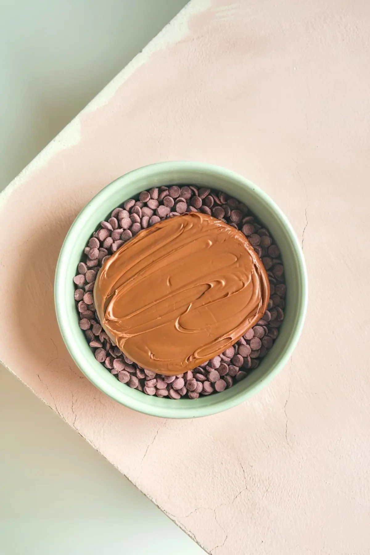 A green bowl filled with chocolate chips topped with a thick layer of melted milk chocolate, placed on a light pink textured surface.
