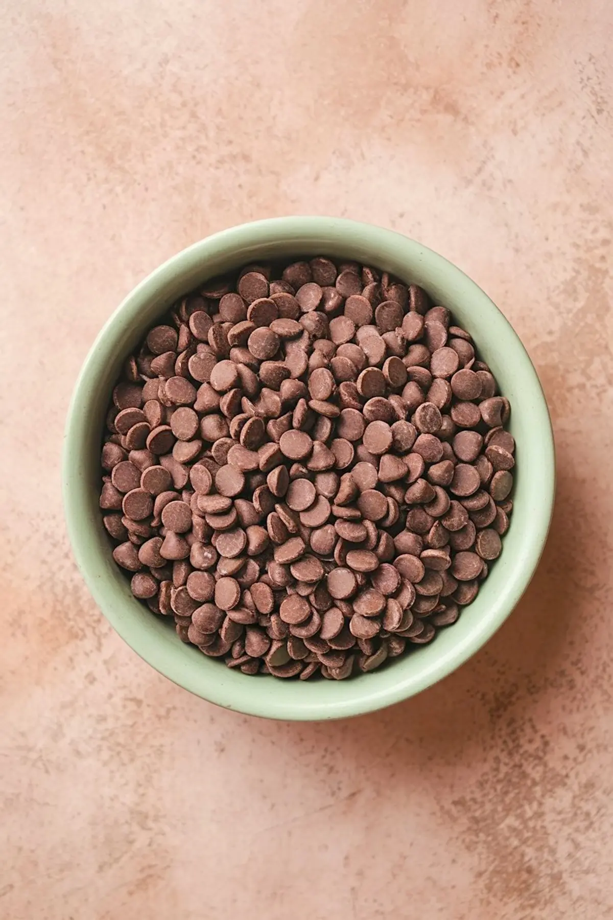 A green bowl filled to the top with semi-sweet chocolate chips placed on a peach-colored textured surface.
