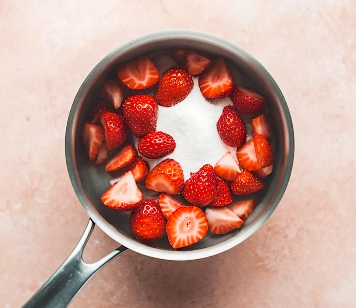 Saucepan containing fresh halved strawberries and a mound of white granulated sugar, ready for cooking, on a soft pink background.