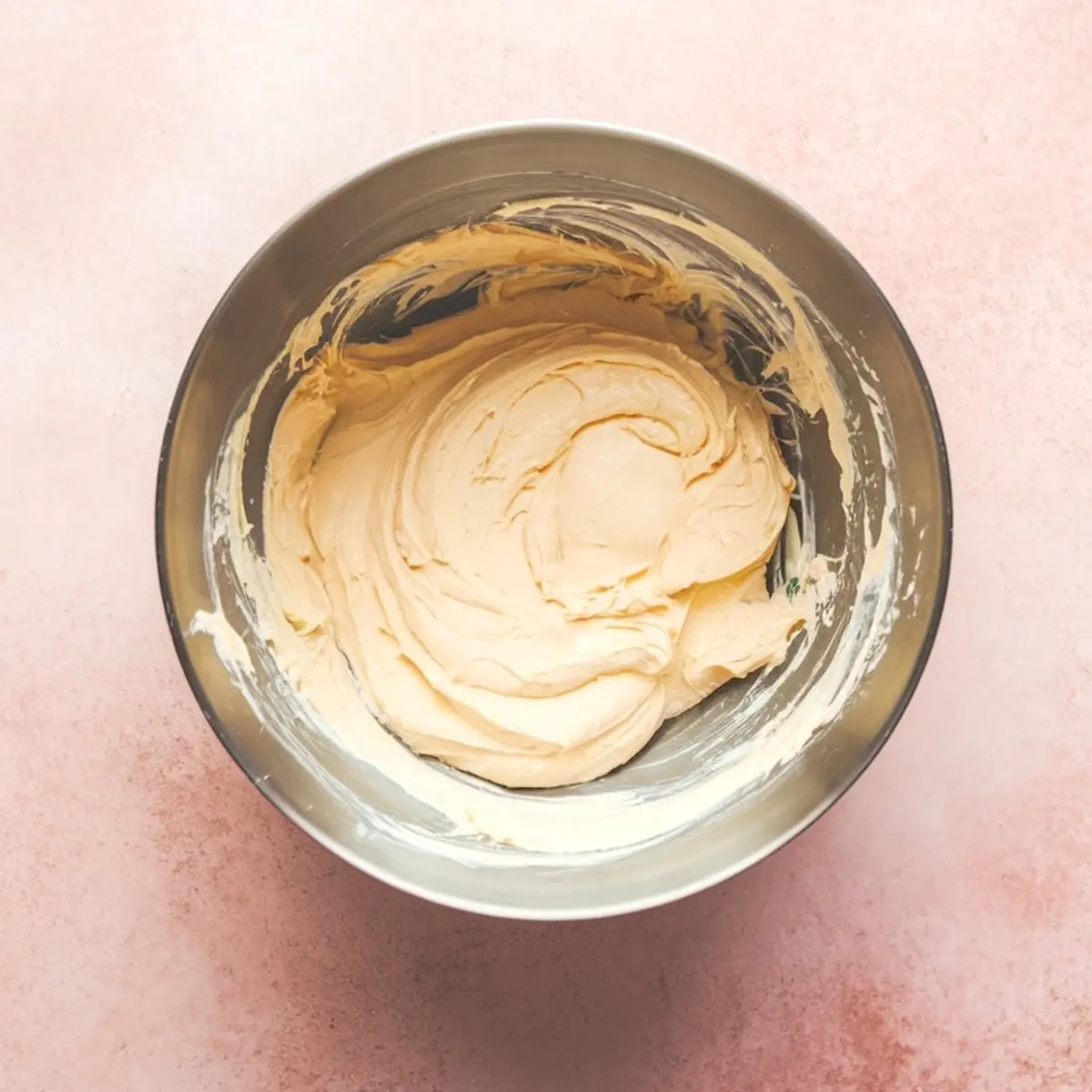 Metal mixing bowl filled with thick, creamy cheesecake mixture after blending, placed on a pink textured surface.