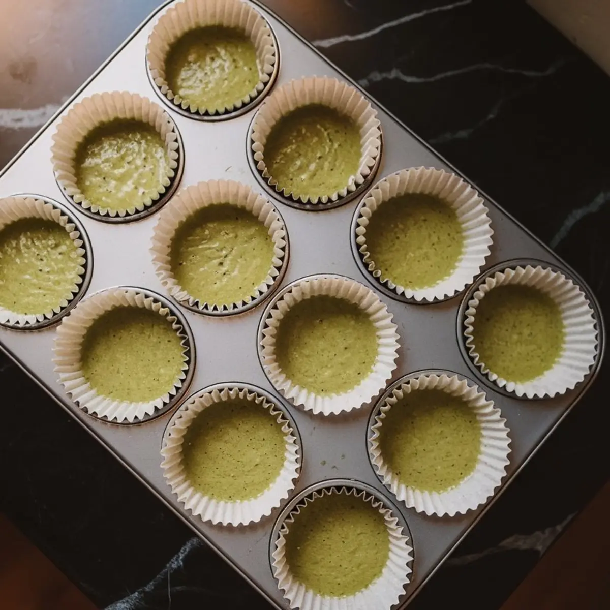 Matcha cupcake batter poured into white paper liners inside a muffin tin, ready for baking on a dark kitchen counter.