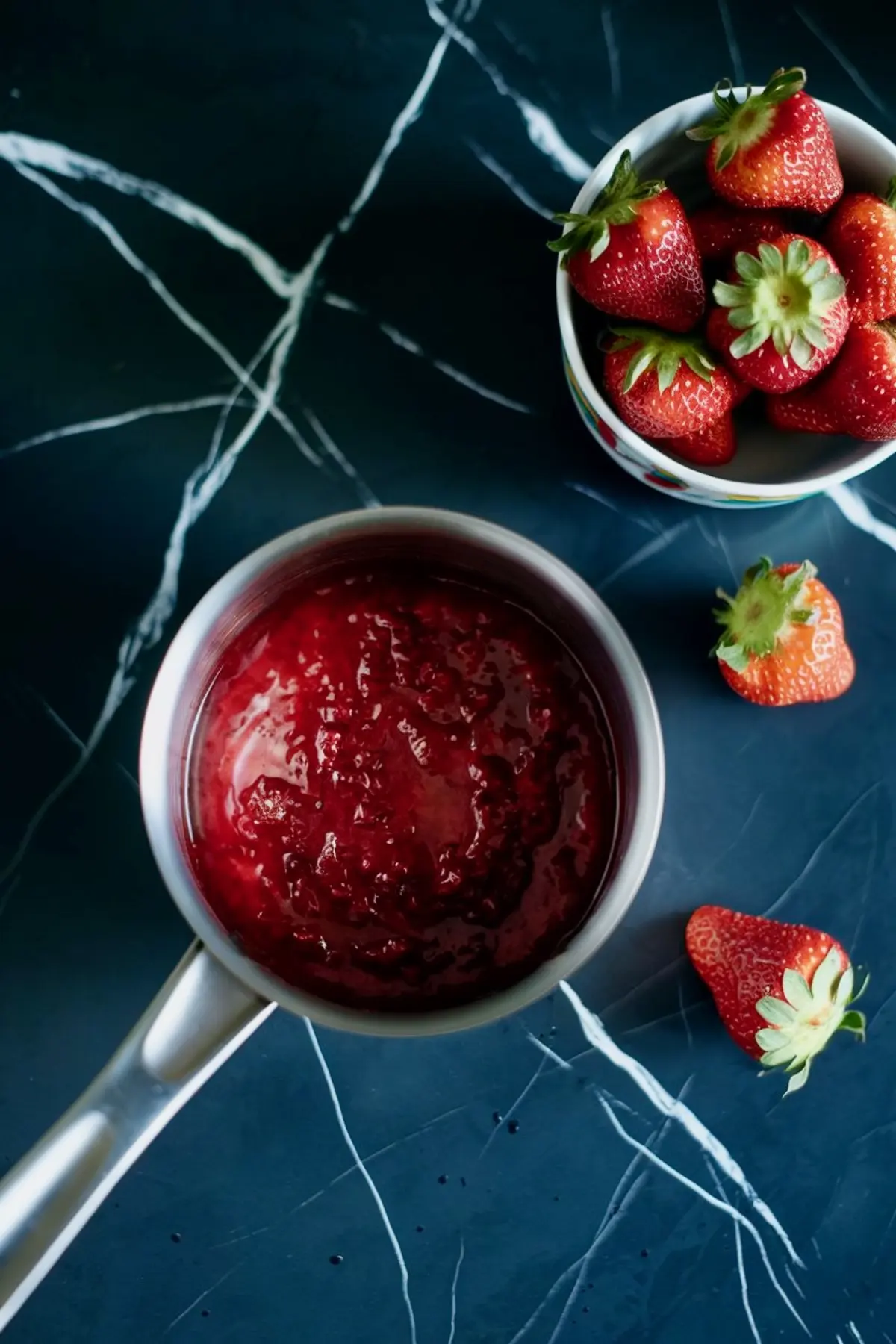 Fresh strawberry compote in a saucepan beside a bowl of ripe strawberries, set on a dark marbled countertop.