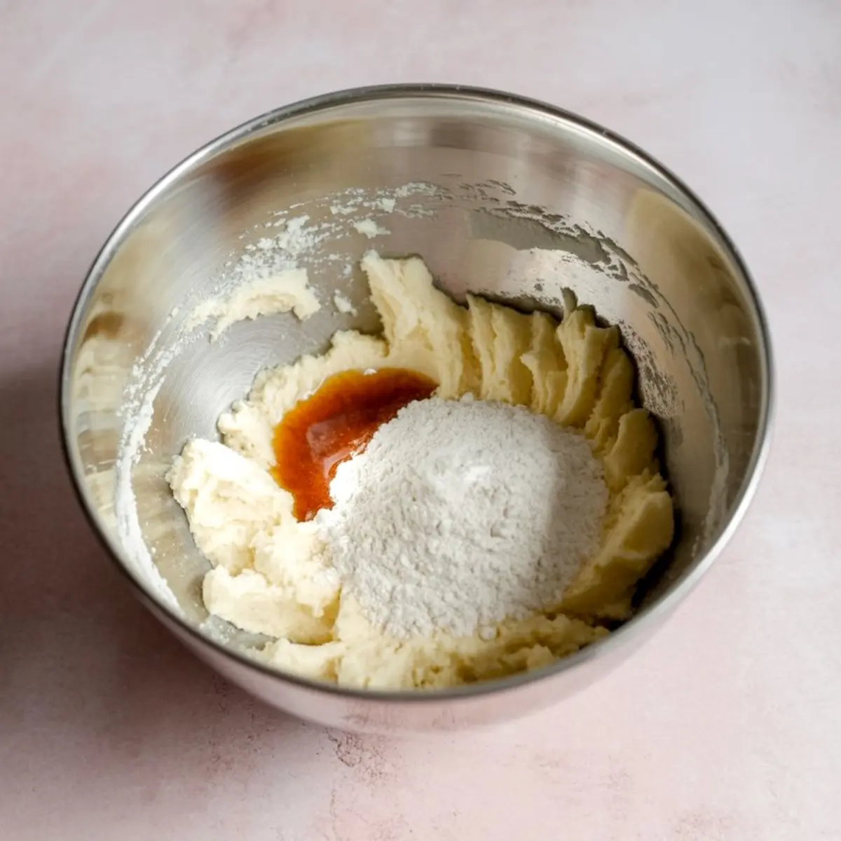 Close-up of a metal mixing bowl containing softened butter, flour, and vanilla extract partially combined for making cookie dough.
