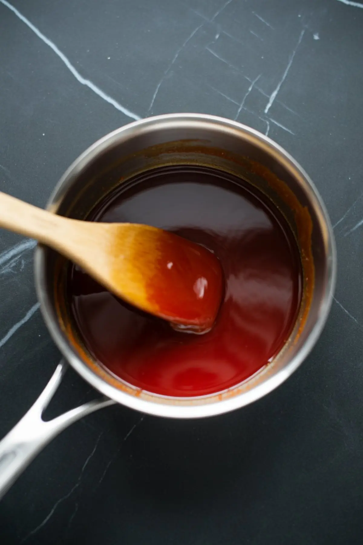 Thick red strawberry glaze being stirred in a saucepan with a wooden spoon on a dark kitchen surface.
