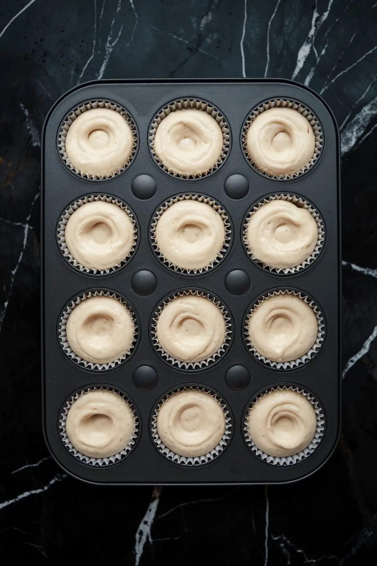 Cupcake pan with twelve cavities filled with smooth vanilla cupcake batter, ready for baking. Evenly portioned batter in white paper liners on a black marble surface, showcasing preparation stage.
