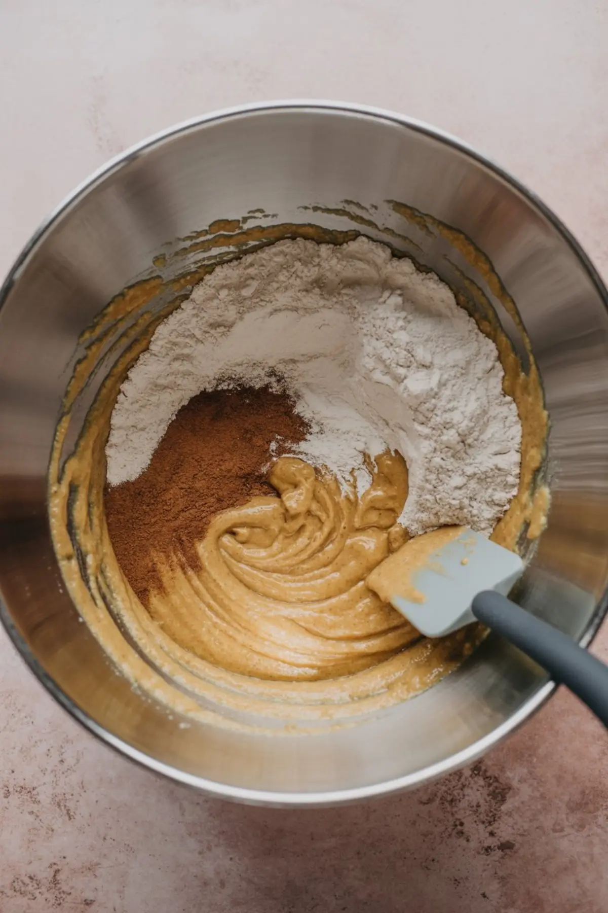 Batter in a mixing bowl with added flour and cinnamon, being stirred with a gray silicone spatula during apple dessert preparation.