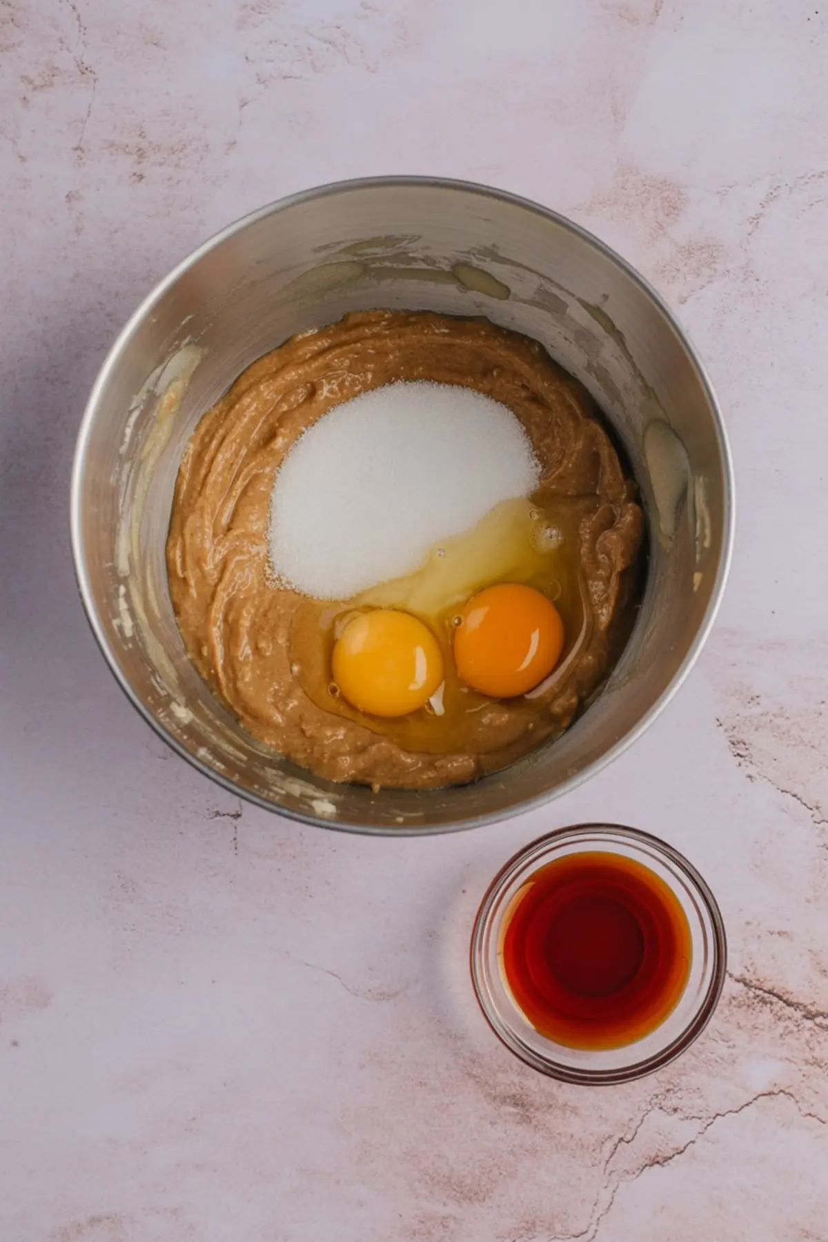 Mixing bowl with thick brown batter, cracked eggs, and granulated sugar on top, next to a small glass bowl of vanilla extract.