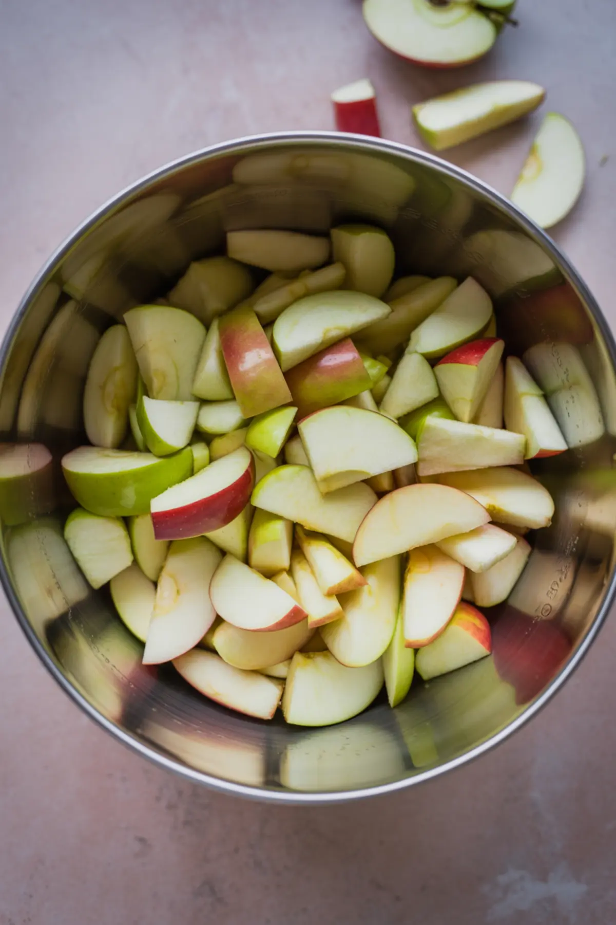 Sliced red and green apples in a stainless steel mixing bowl on a light kitchen counter, prepared for baking or cooking.