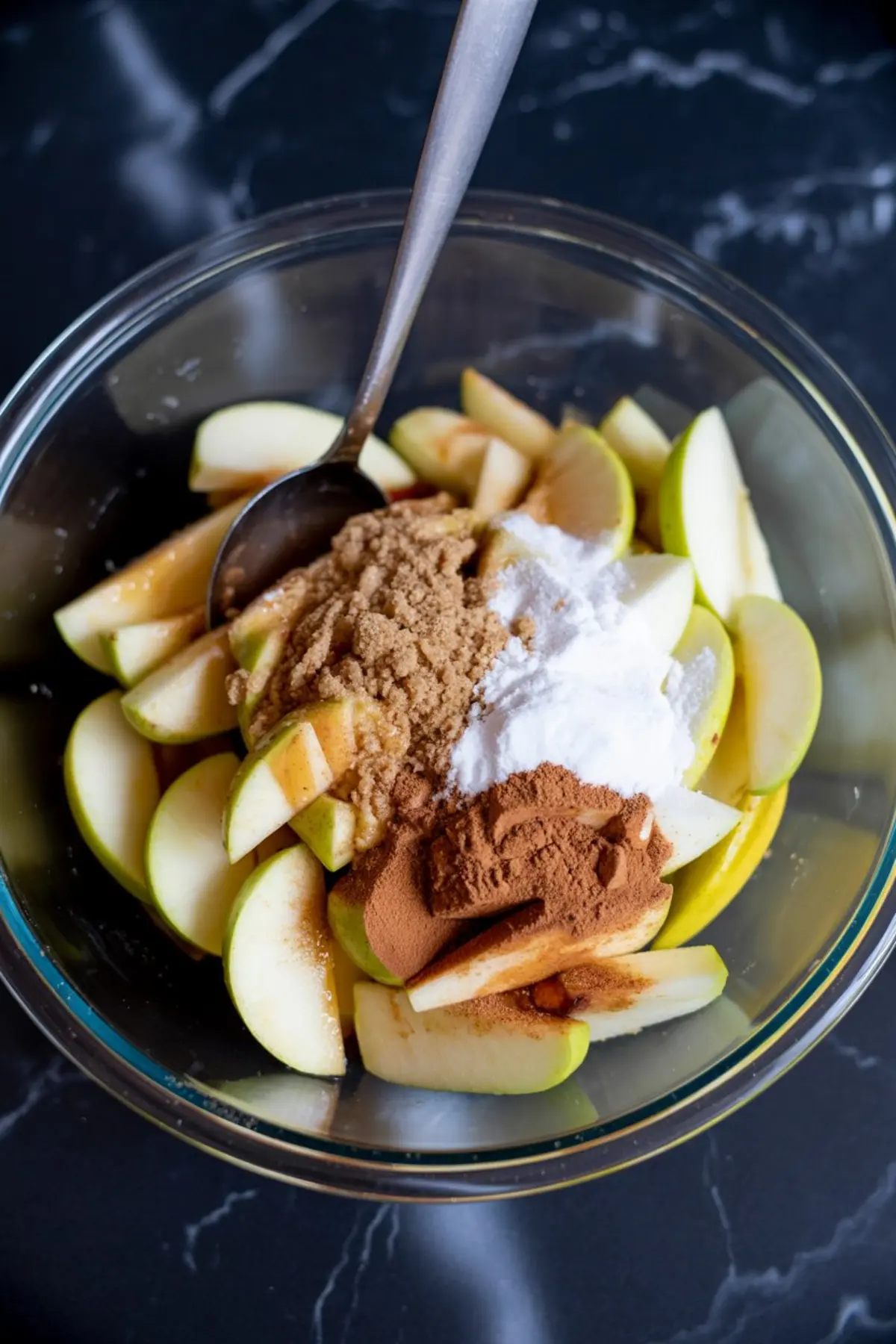 Glass bowl filled with sliced green apples topped with brown sugar, white sugar, ground cinnamon, and flour on a dark marble countertop, ready for apple pie filling.