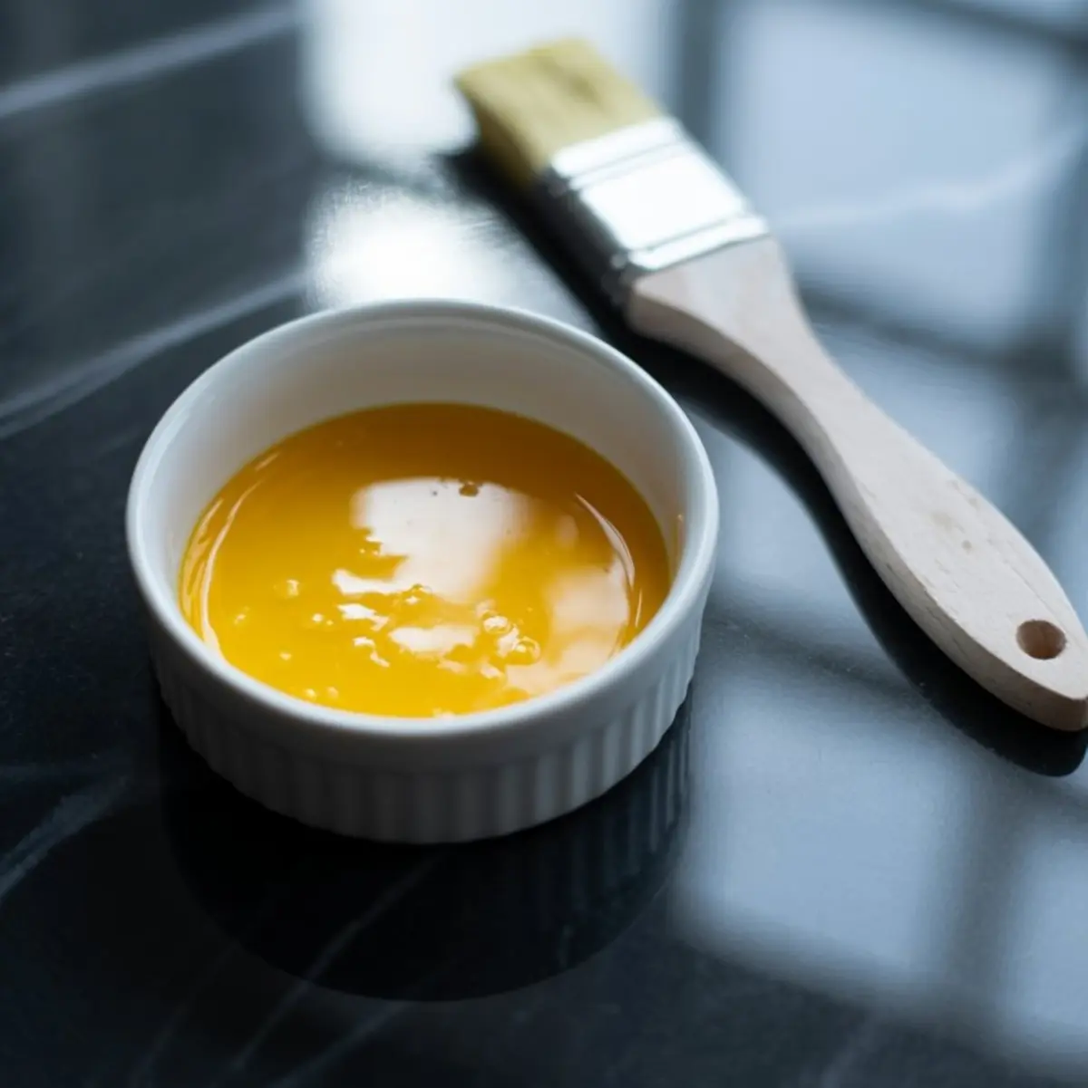 Small white ramekin filled with glossy egg wash next to a pastry brush on a black countertop, commonly used for adding golden shine to pie crusts and baked goods.