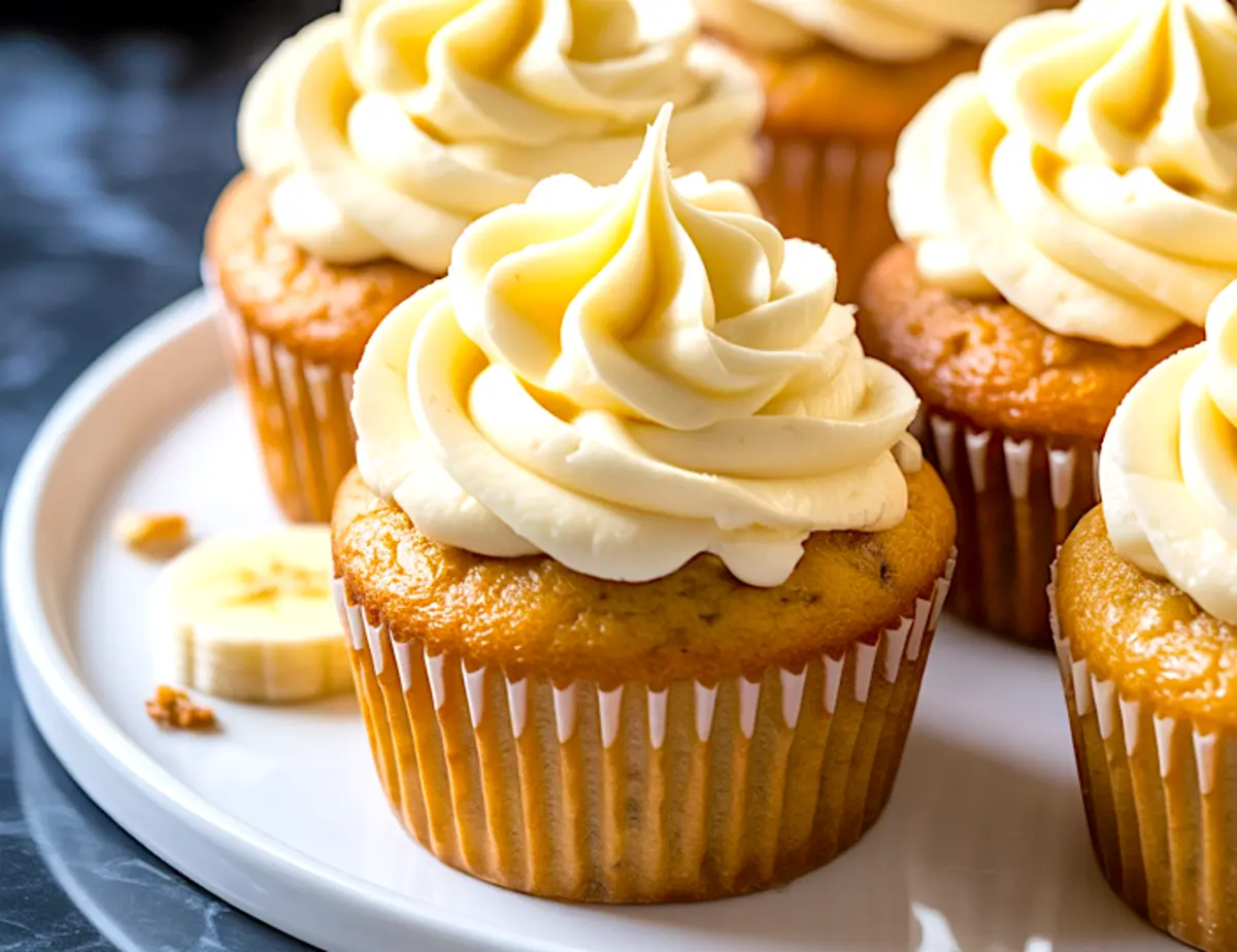 Frosted banana cupcakes displayed on a white plate, topped with creamy swirls of vanilla frosting and garnished with banana slices.