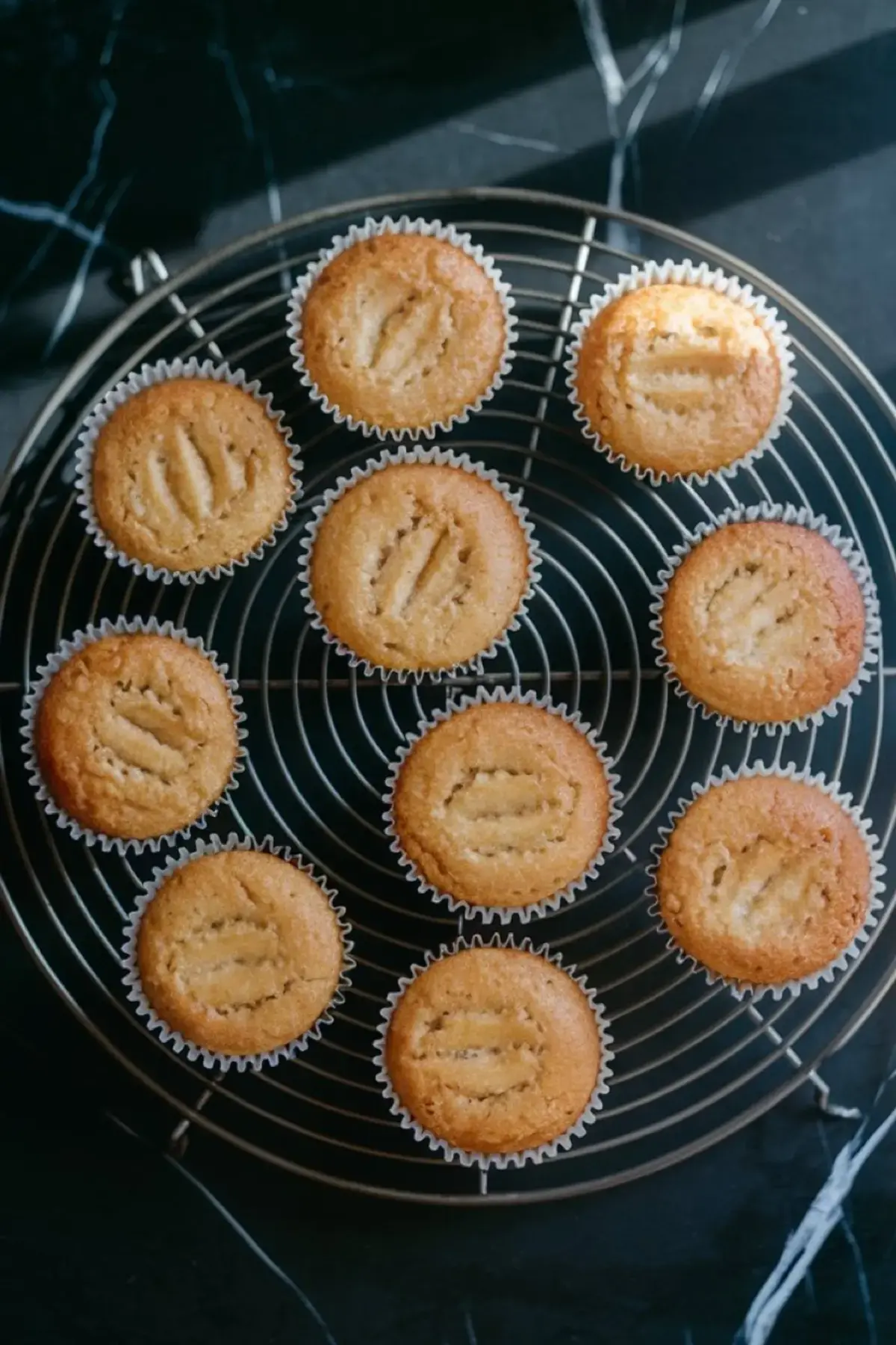 Freshly baked banana cupcakes with golden tops cooling on a round wire rack, each topped with baked-in banana slices.