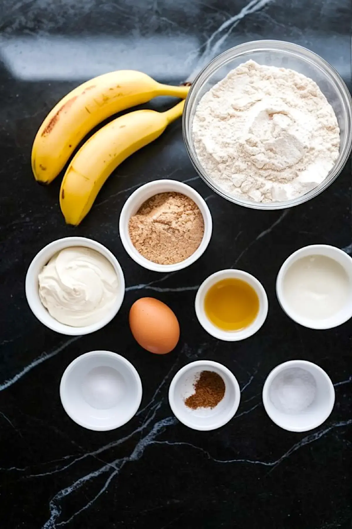 Overhead view of banana cupcake ingredients, including ripe bananas, all-purpose flour, brown sugar, sour cream, egg, oil, vanilla, baking soda, baking powder, and cinnamon on a black marble background.