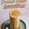 Tall glass of banana peanut butter smoothie on a blue plate, topped with chopped peanuts and a swirl of peanut butter, surrounded by scattered crushed peanuts and a silver spoon on a light marble surface with large text above.