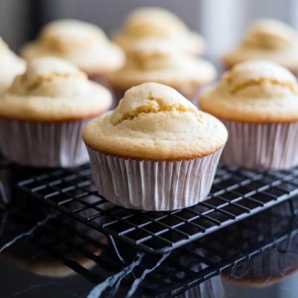 Golden vanilla cupcakes in white liners cooling on a black wire rack, with puffed tops and a soft crumb texture visible.