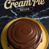 Whole Boston cream pie covered in shiny chocolate ganache, displayed on a yellow plate over a black marble surface. The text “Boston Cream Pie Recipe” appears above the cake.