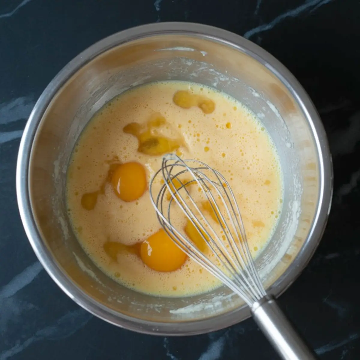 Unmixed eggs sitting on top of a yellow batter in a stainless steel bowl with a metal whisk. The mixture is just beginning to combine.
