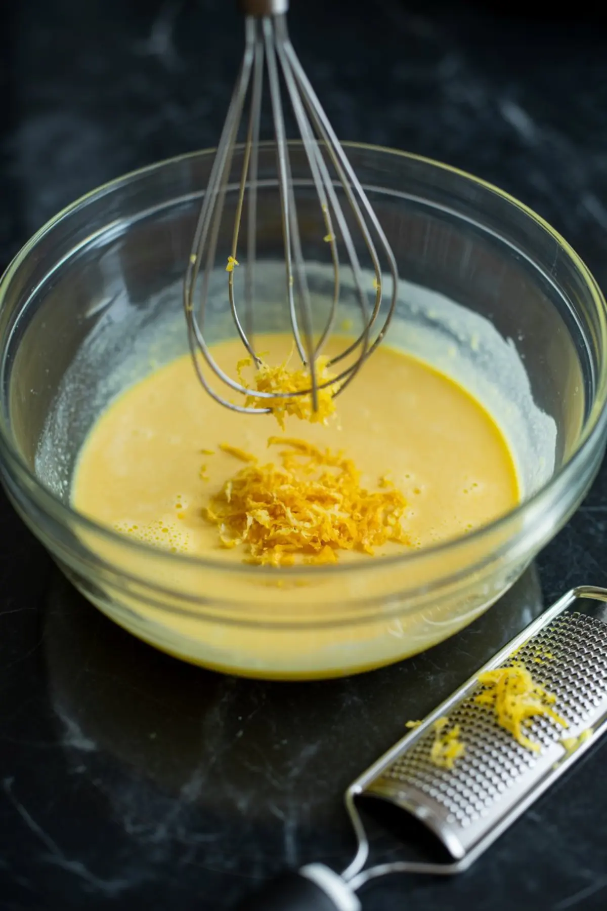 Fresh lemon zest falling into a bowl of yellow liquid batter with a whisk and microplane zester on a dark marble counter.