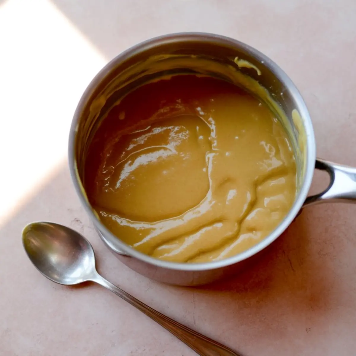 Saucepan of thickened butterscotch pudding filling with a glossy surface, placed beside a metal spoon on a light pink surface.