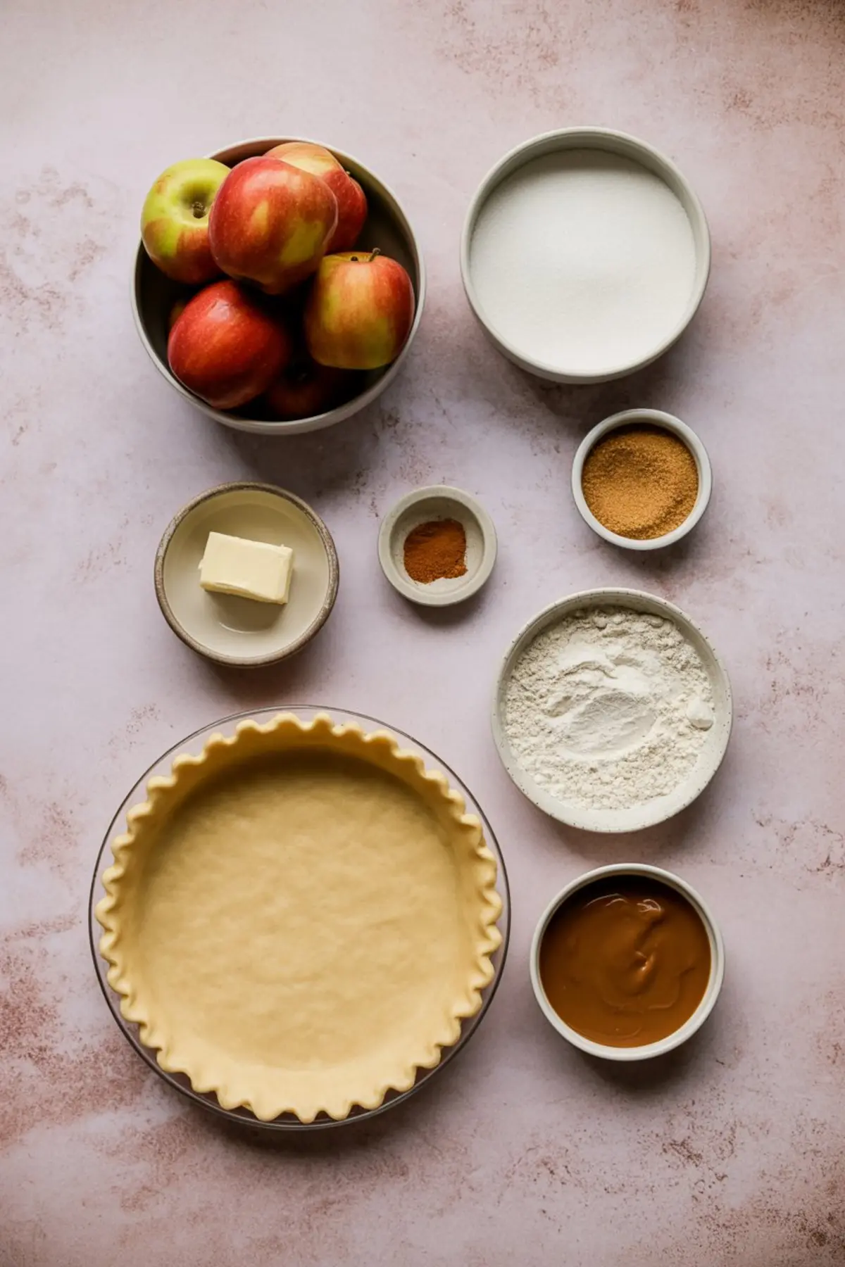Flat lay of caramel apple pie ingredients on a blush-toned surface, including whole apples, granulated sugar, brown sugar, butter, cinnamon, flour, caramel sauce, and an unbaked pie crust.