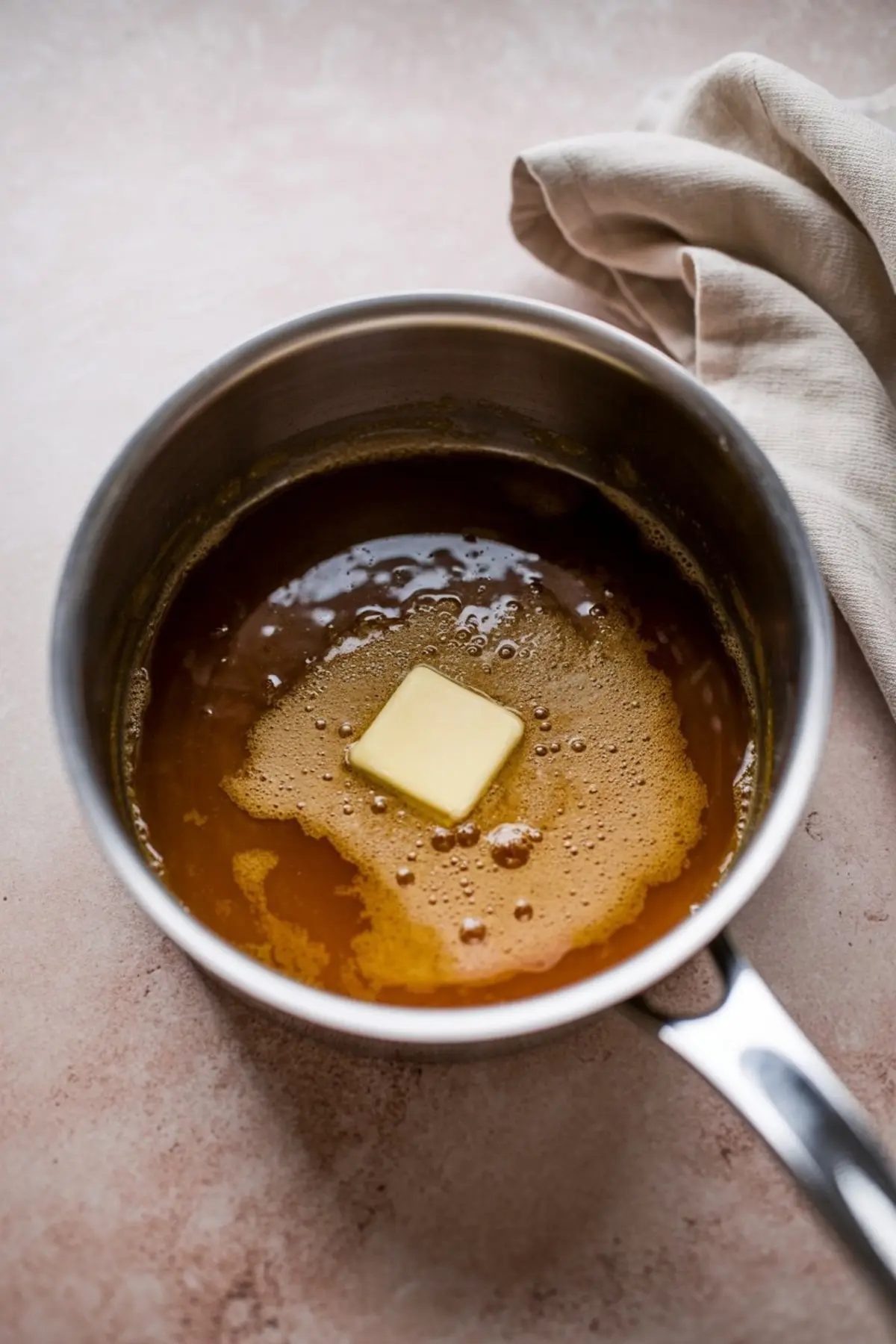 Overhead view of a stainless steel saucepan with caramelized butter mixture bubbling on a light surface, featuring a square pat of butter slowly melting in the center.