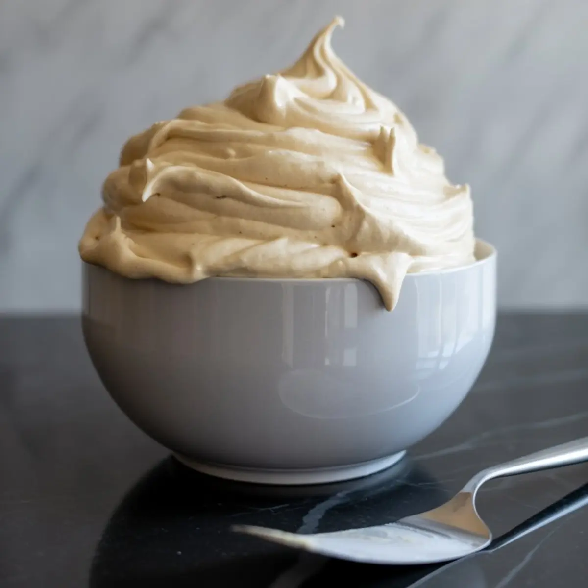 Fluffy cinnamon cream cheese frosting piled high in a white ceramic bowl, with a spatula beside it on a glossy black marble surface.