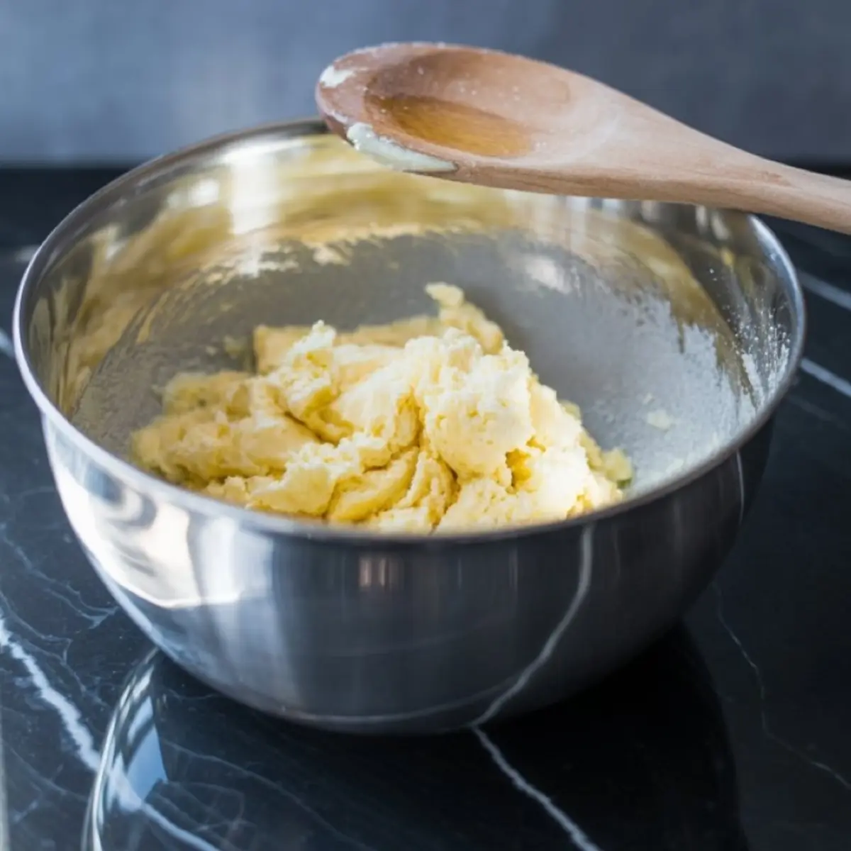 Thick, coarse dough mixture in a metal bowl with a wooden spoon coated in batter, set on a black marble kitchen counter.