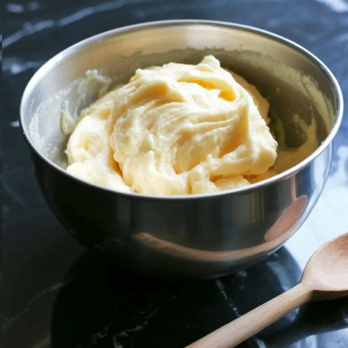 Smooth and fluffy cake batter in a stainless steel mixing bowl placed on a dark marble surface next to a wooden spoon.