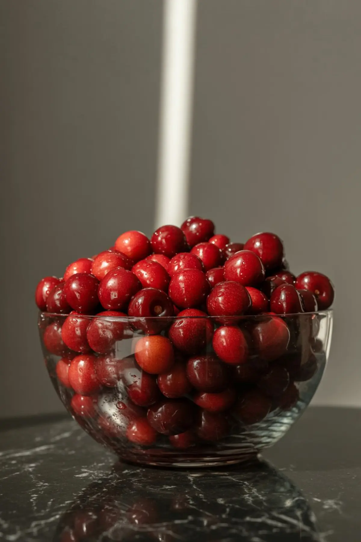 Clear glass bowl filled with shiny red cherries without stems, stacked high on a dark marble countertop under warm lighting.