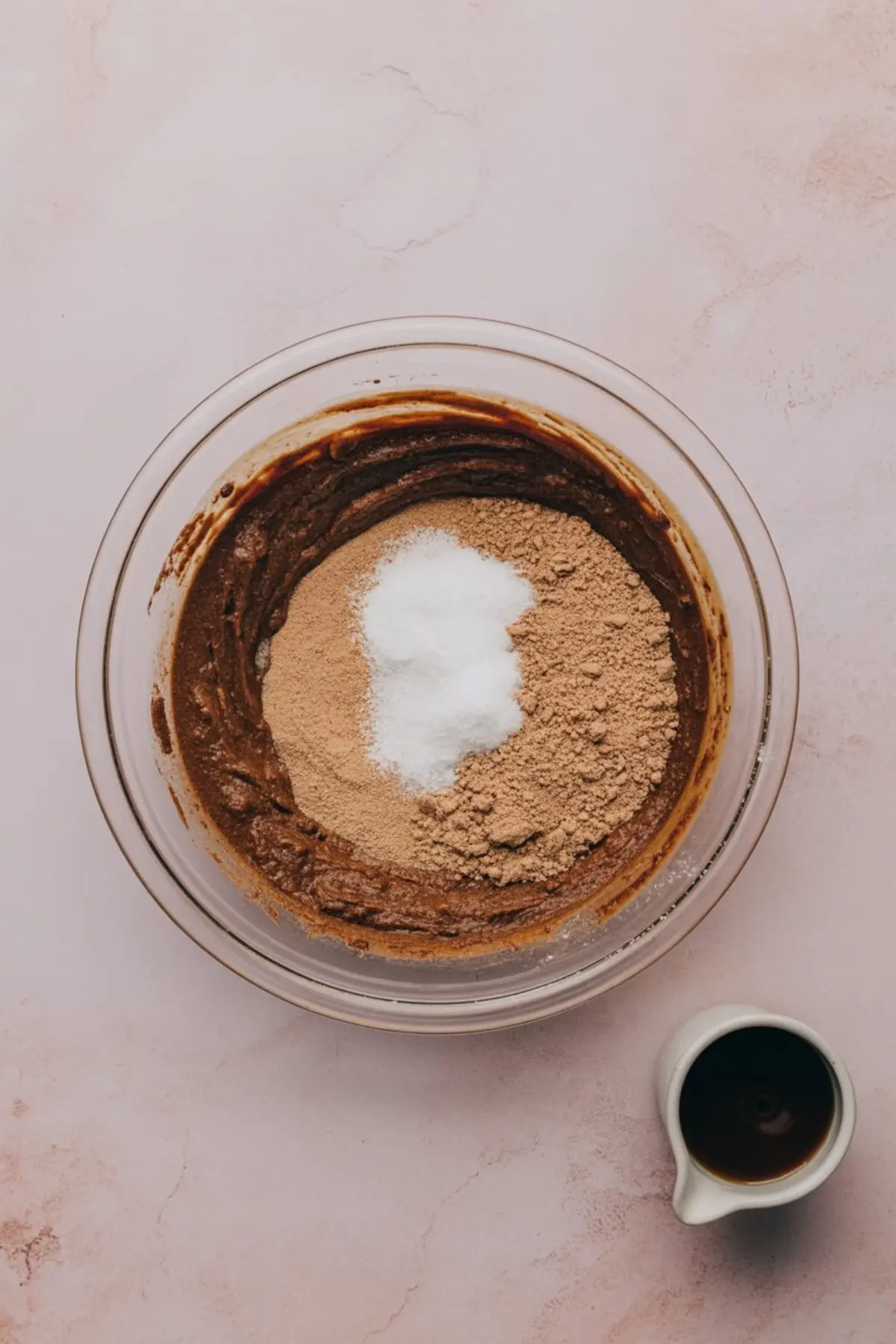 Glass mixing bowl filled with chocolate batter, topped with brown sugar and white granulated sugar, placed on a pink surface next to a small white jug of dark liquid.