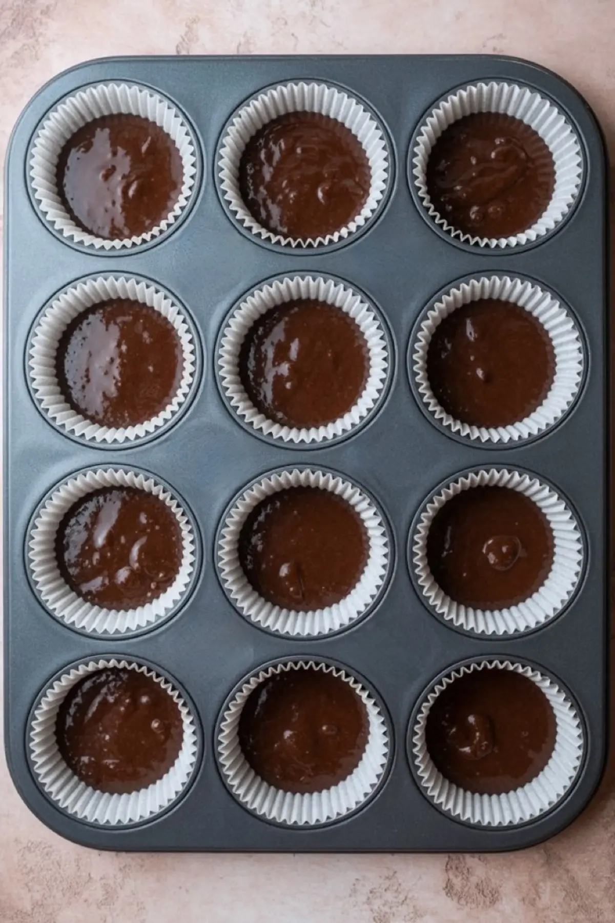 Muffin tin filled with chocolate cupcake batter in white paper liners, ready for baking on a light pink surface.