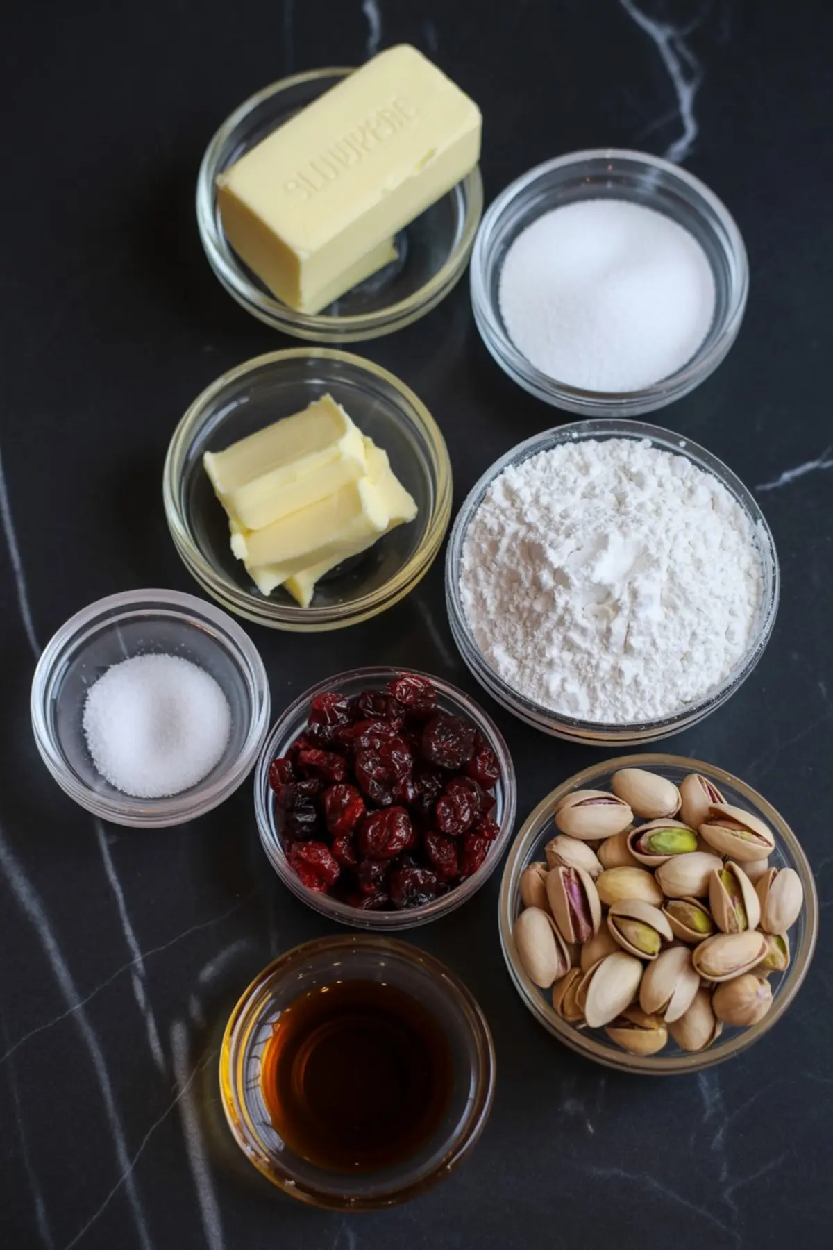 Overhead view of ingredients for cranberry pistachio shortbread cookies, including butter, sugar, flour, salt, dried cranberries, pistachios, and vanilla extract arranged on a black marble surface.