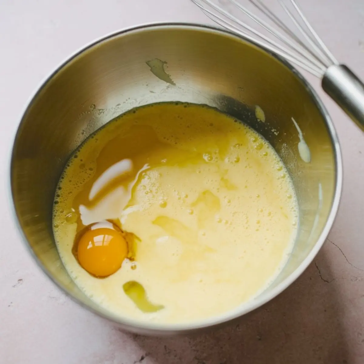 Stainless-steel bowl shows frothy egg-and-butter mixture with one intact yolk; a metal whisk rests on the rim over a blush countertop, capturing early Dutch apple-pie filling prep, home-baking, fall-dessert vibes.