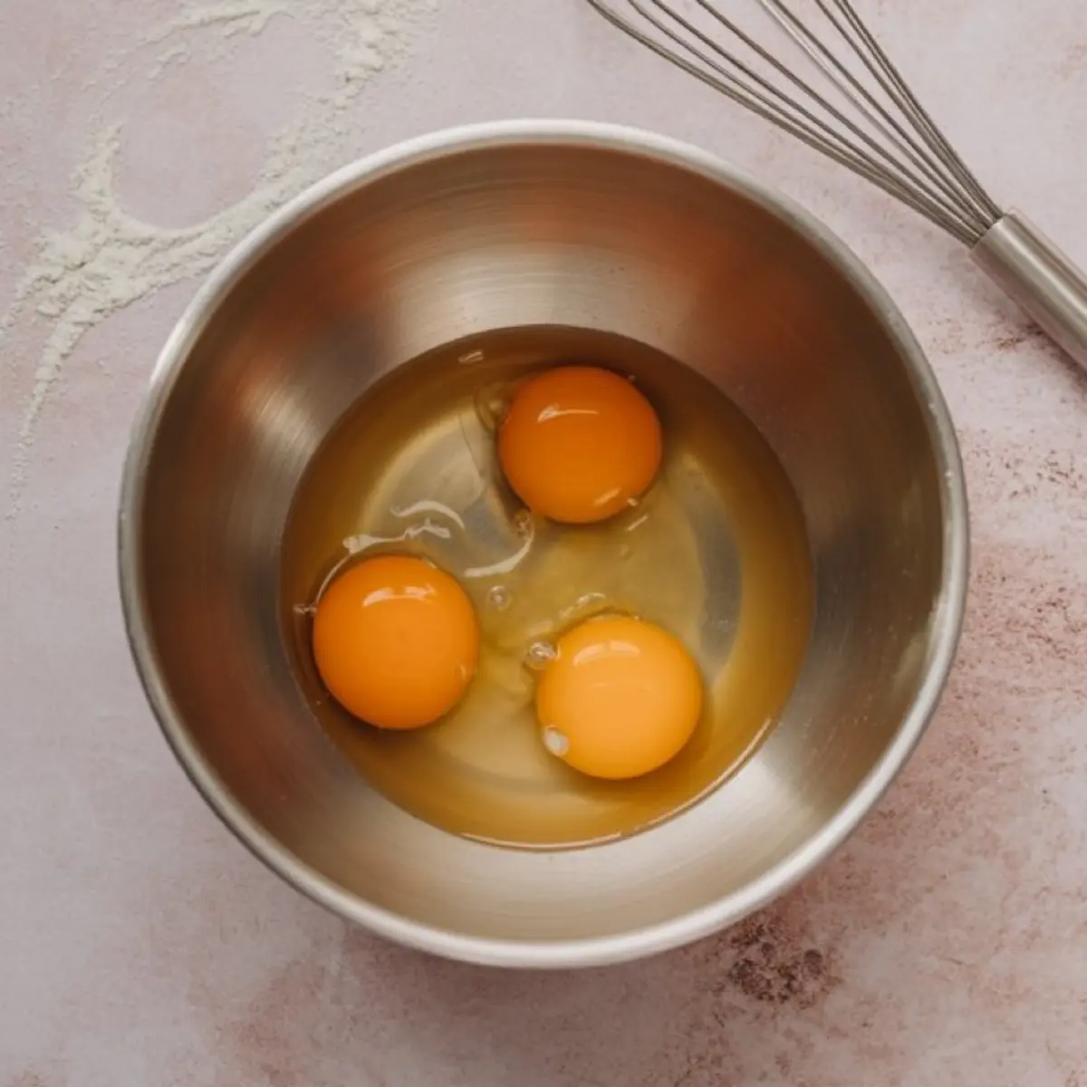 Three cracked raw eggs in a stainless steel bowl, placed on a floured pink surface next to a metal whisk, ready for baking.
