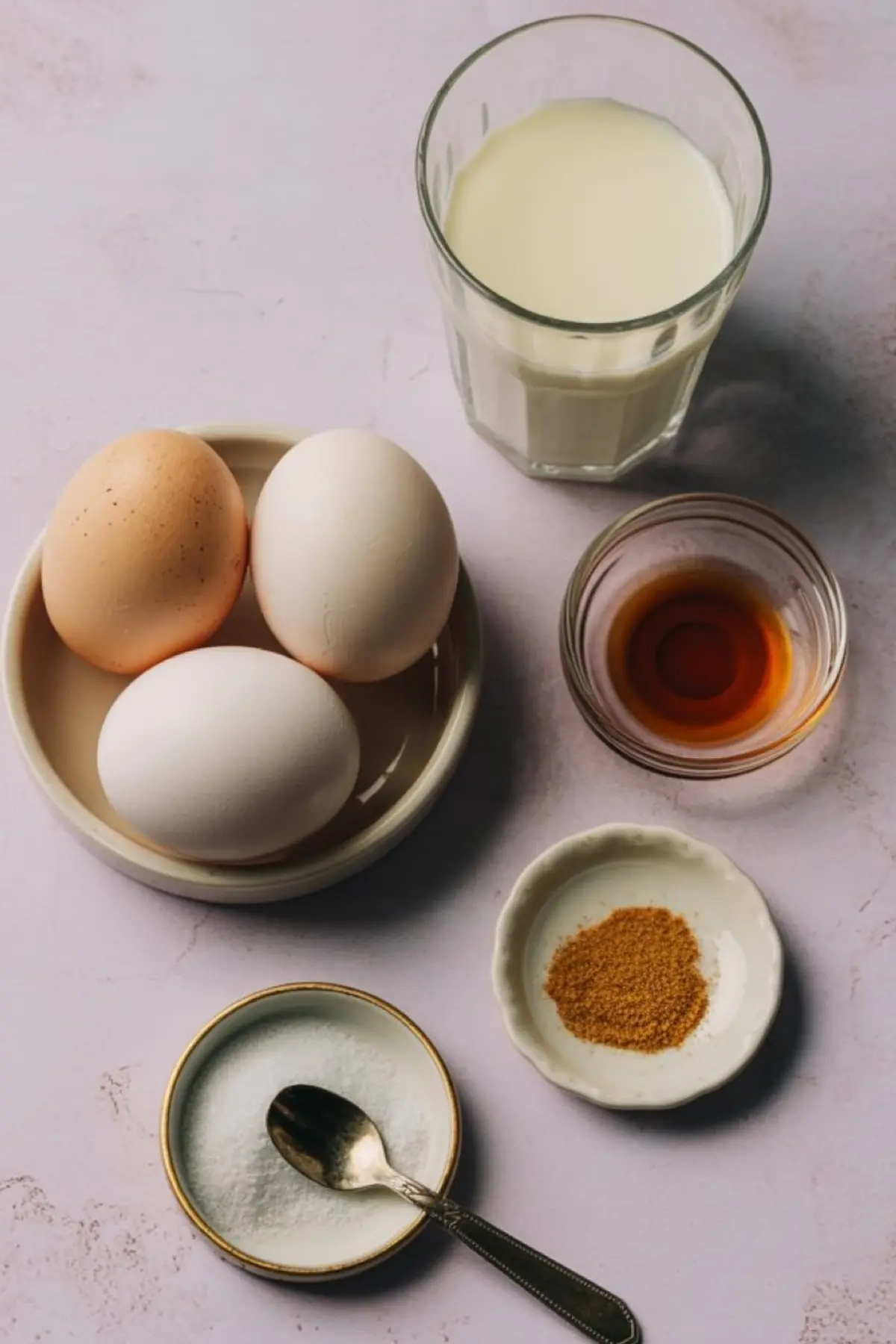 Flat lay of egg custard pie ingredients including eggs, milk, vanilla extract, granulated sugar, and ground nutmeg on a light pink background.
