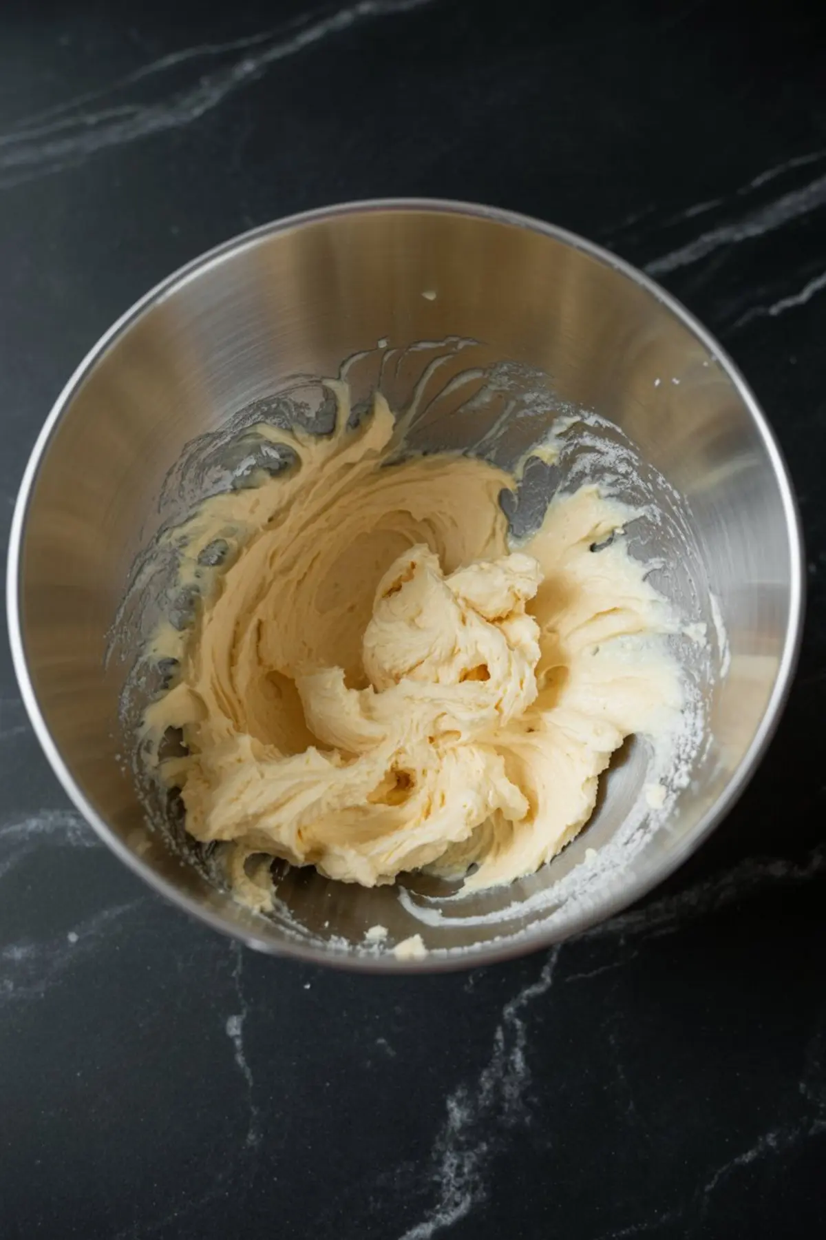 Metal mixing bowl with whipped butter and sugar creamed into a light, fluffy mixture sitting on a black marble counter.