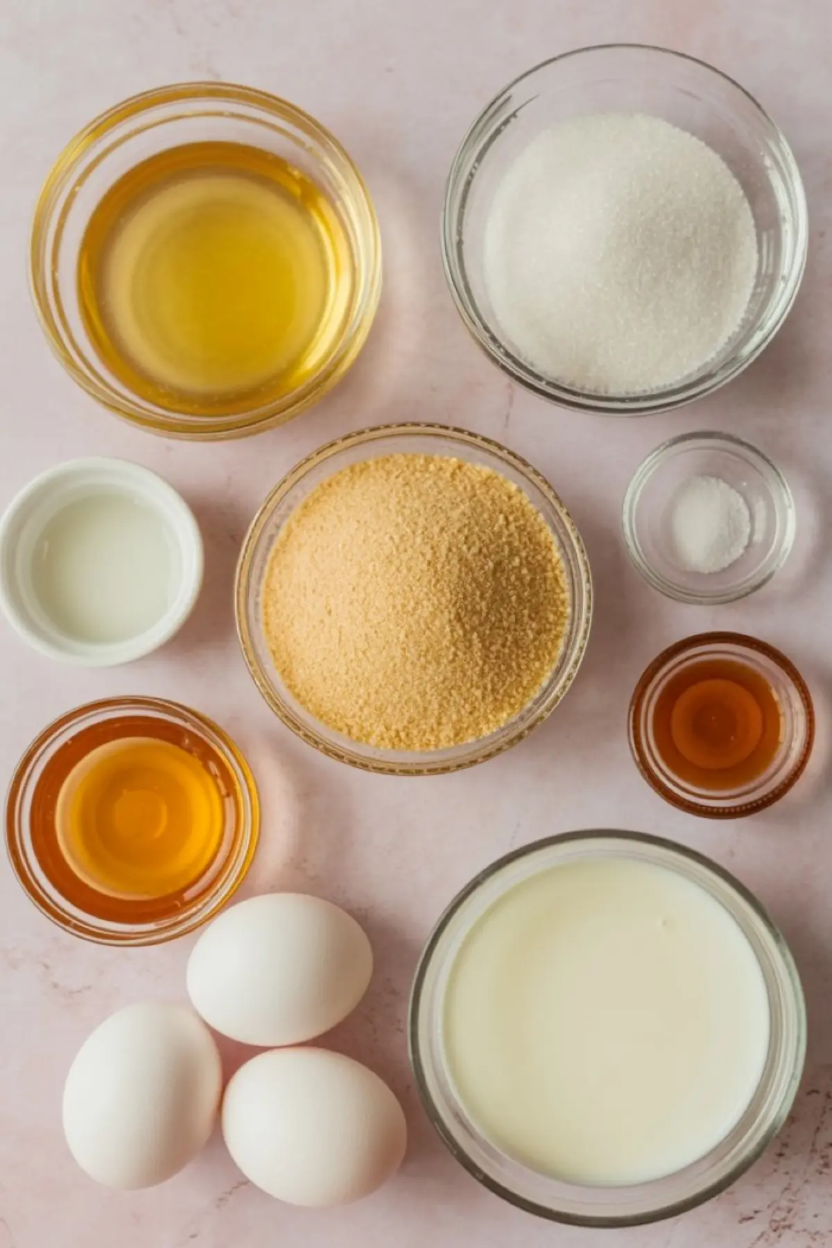 Flat-lay of honey-pie ingredients arranged neatly on a soft-pink background: glass bowls of golden honey, white sugar, graham-cracker crumbs, milk, vegetable oil, vanilla extract, and salt, plus three white eggs ready for baking.