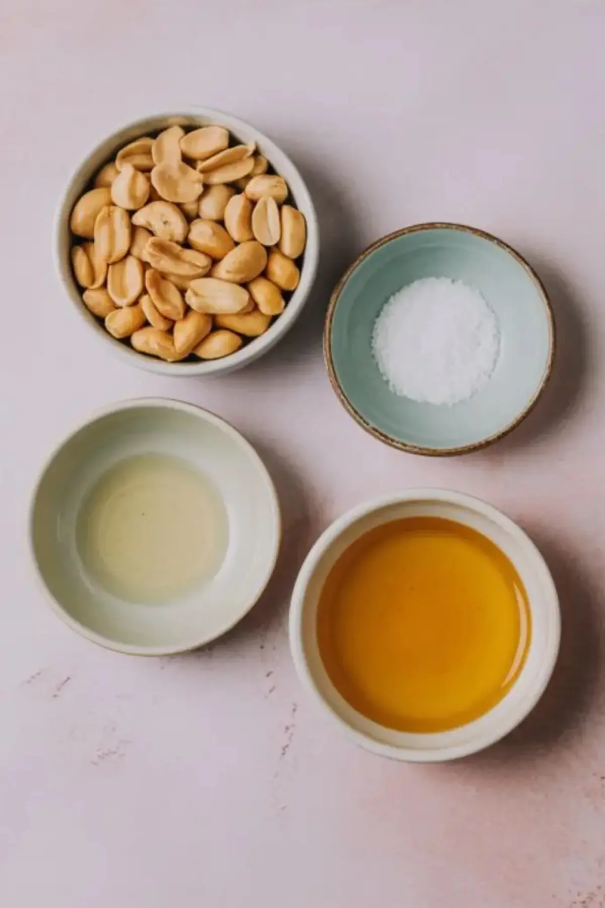 Flat lay of small bowls filled with raw peanuts, sea salt, honey, and oil on a light pink background, showcasing ingredients for homemade peanut butter.