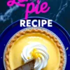Overhead view of a whole lemon pie with whipped topping and lemon zest, placed in a foil pie dish on a dark marble surface with one slice removed.