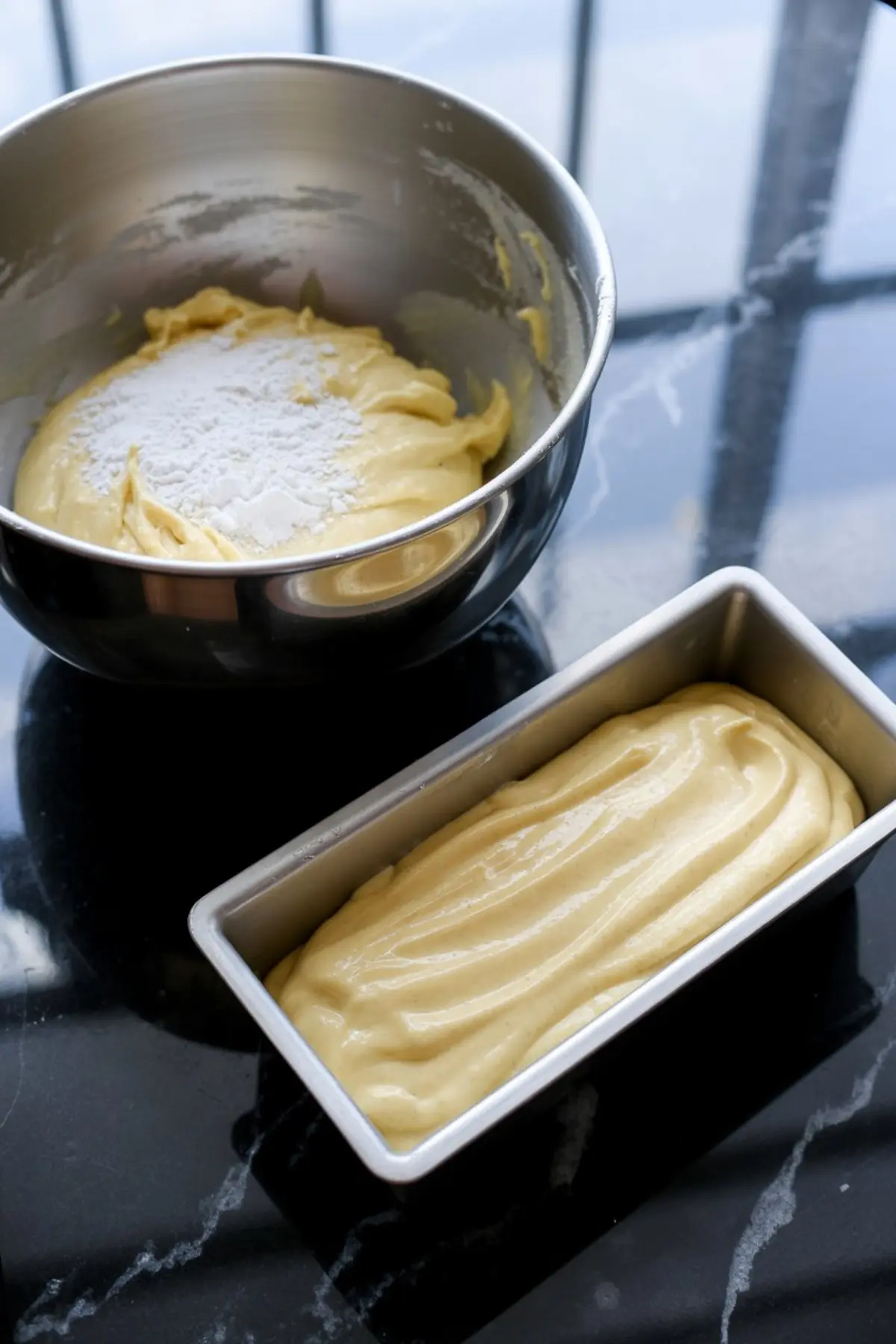 Cake batter in a metal mixing bowl with flour on top, next to a loaf pan filled with smoothed batter, ready for baking on a dark stone surface.
