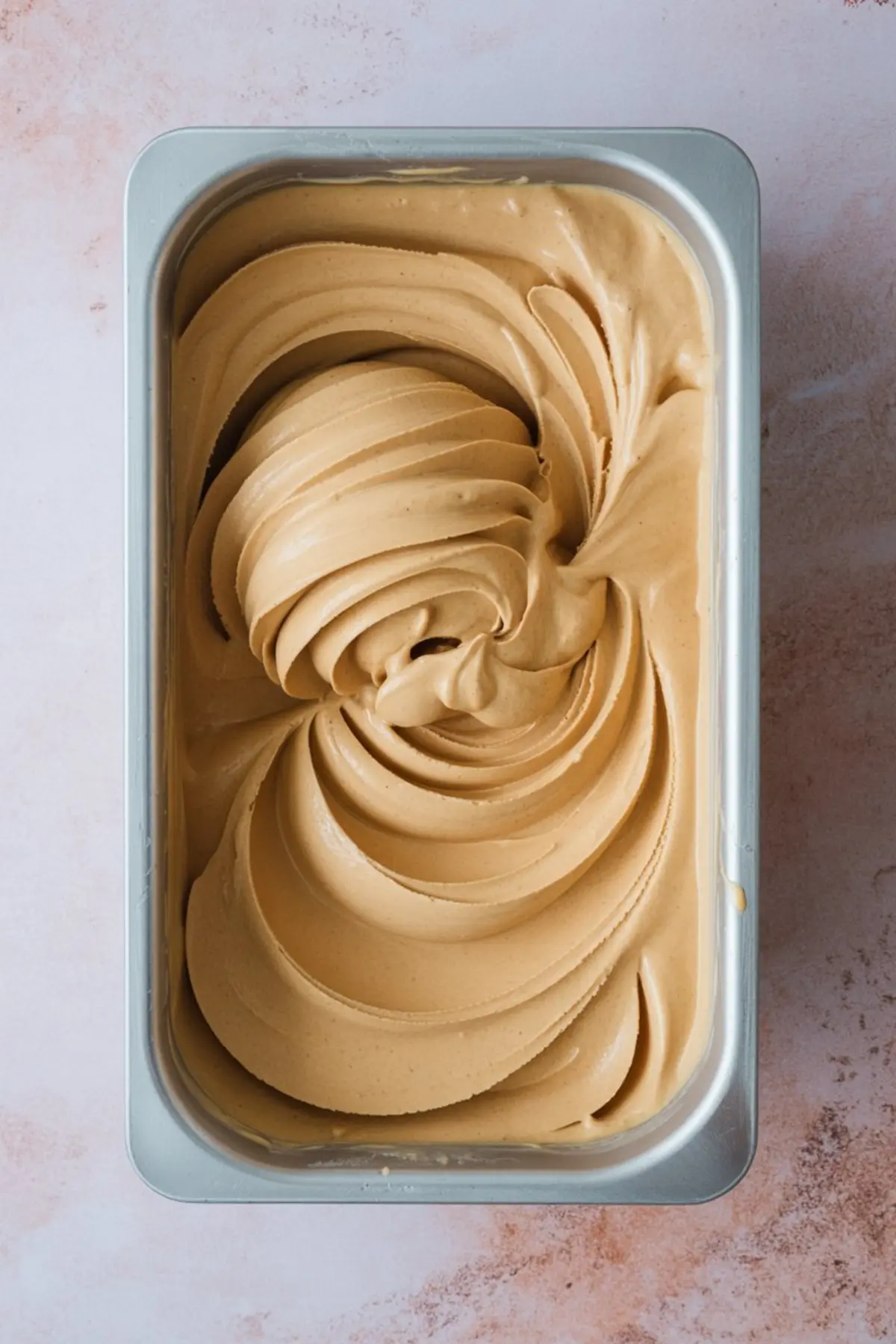 Creamy peanut butter banana ice cream swirled into a metal container, showing smooth texture and rich caramel color, ideal for frozen dessert recipes and summer treat inspiration.