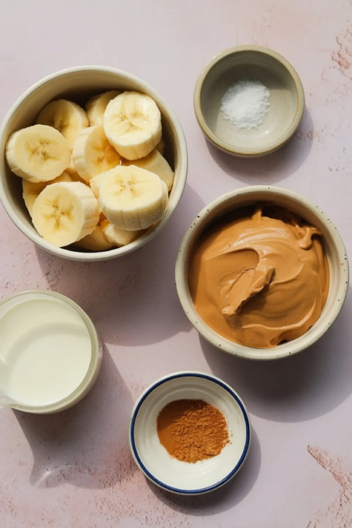 Flat lay of peanut butter banana ice cream ingredients including sliced bananas, creamy peanut butter, milk, ground cinnamon, and sea salt in ceramic bowls on a pastel pink background.