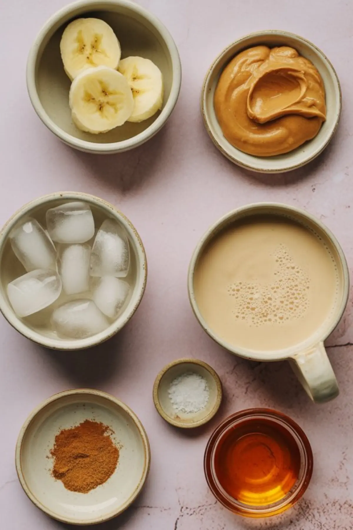 Flat lay of banana smoothie ingredients on a pink background, including sliced bananas, creamy peanut butter, ice cubes, brewed coffee or milk, ground cinnamon, vanilla extract, and a pinch of salt in small ceramic bowls.