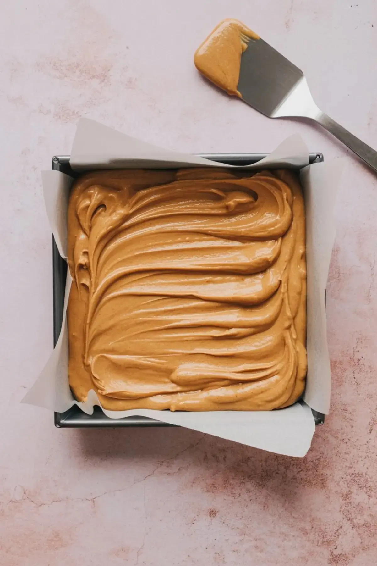Baking pan lined with parchment paper filled with smooth peanut butter mixture spread evenly with a spatula on the side.
